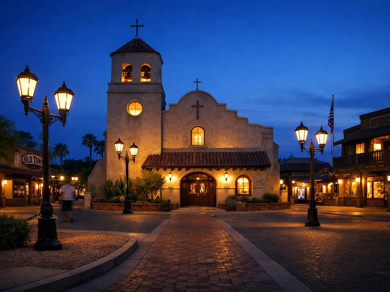 A lit stucco church with a bell tower stands in a quiet plaza at dusk, flanked by streetlamps and shops